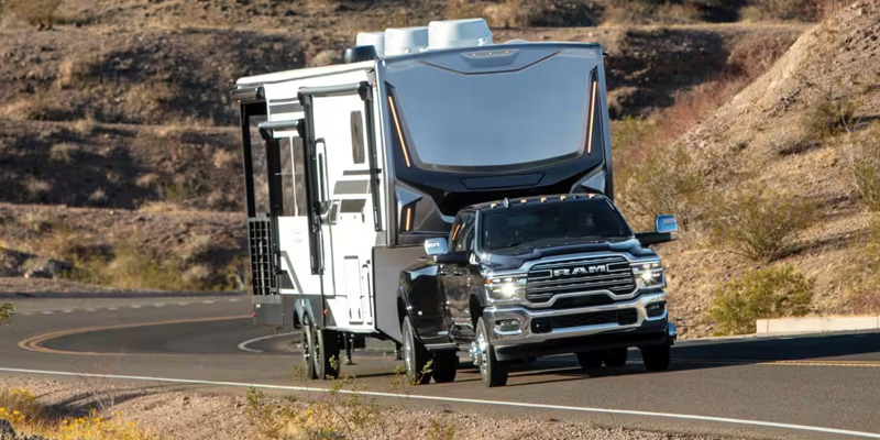 A RAM truck towing a large RV trailer along a curved road in a desert landscape.